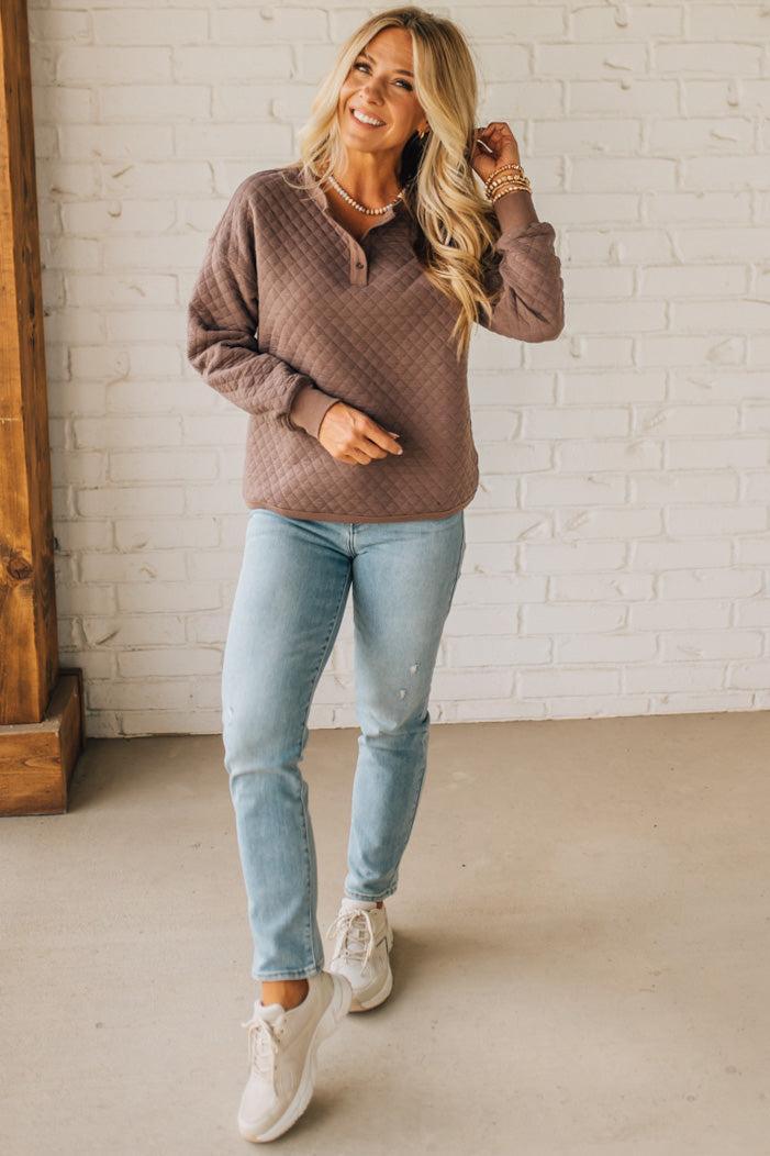 Woman wearing a brown sweater, light blue jeans, and white sneakers in a room with a white brick wall.
