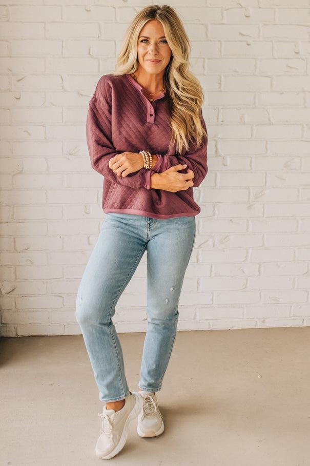 Woman wearing a maroon hoodie and light blue jeans against a white brick wall.