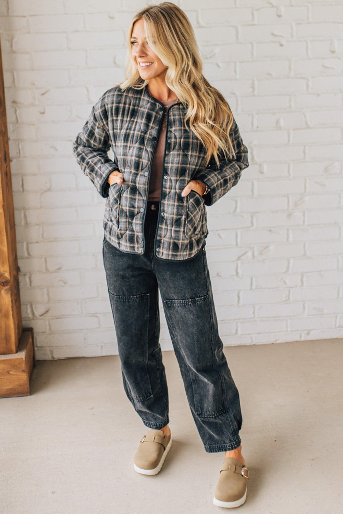 Woman wearing a plaid jacket and dark jeans standing against a white brick wall.