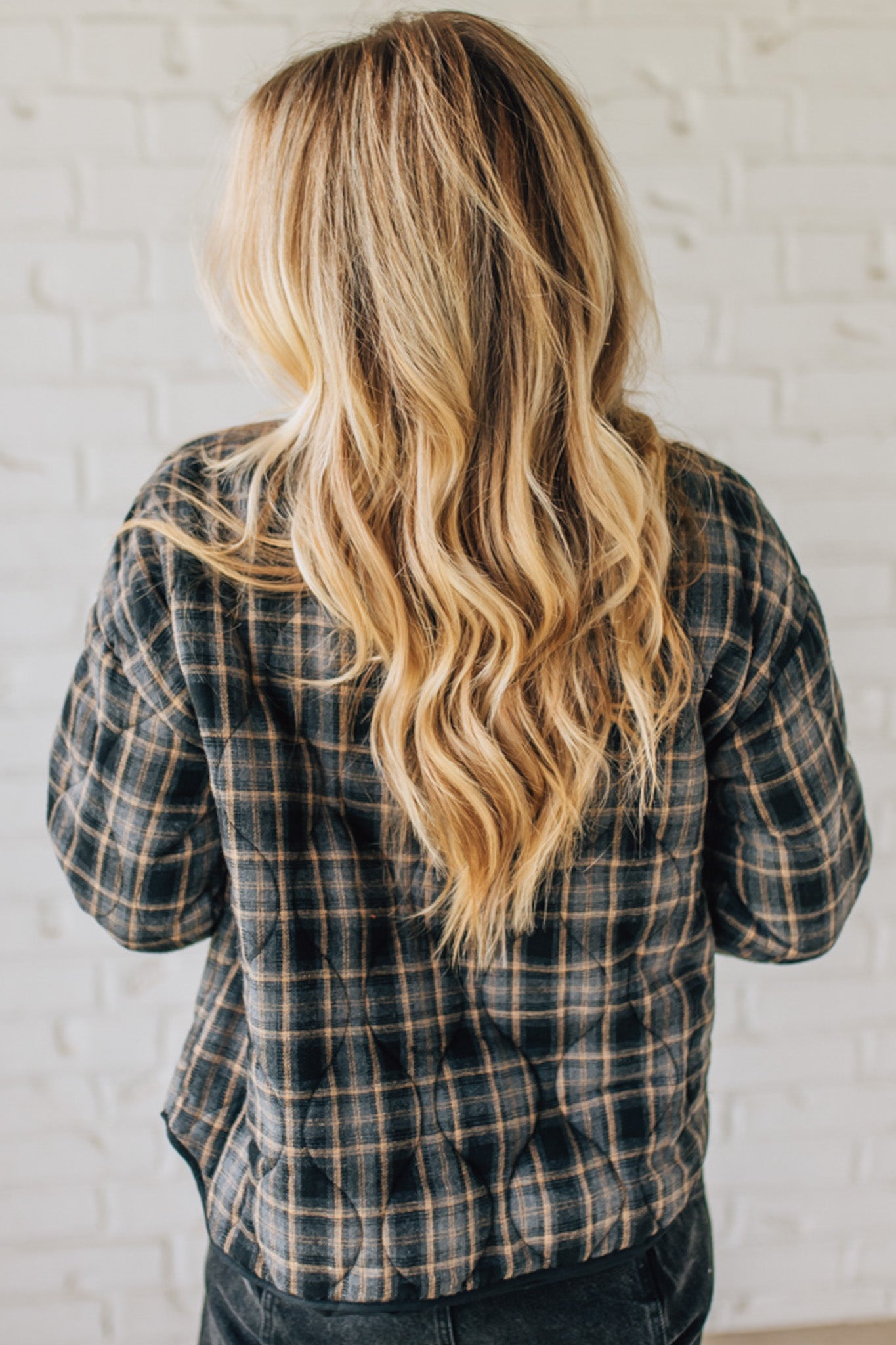 Person wearing a plaid shirt with long blonde hair against a white brick wall.