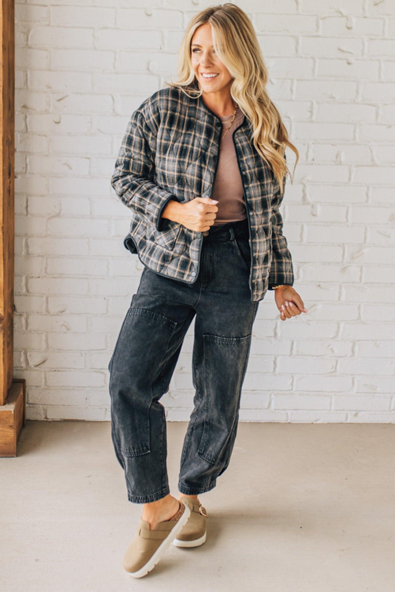 Woman wearing a plaid shirt and dark pants standing against a white brick wall.