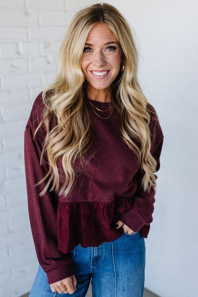 Woman wearing a burgundy sweater and blue jeans against a white brick wall.