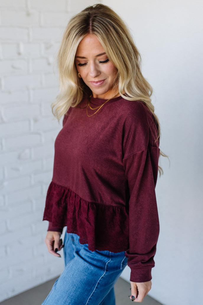 Woman wearing a burgundy long-sleeve top with ruffled details against a white brick wall.