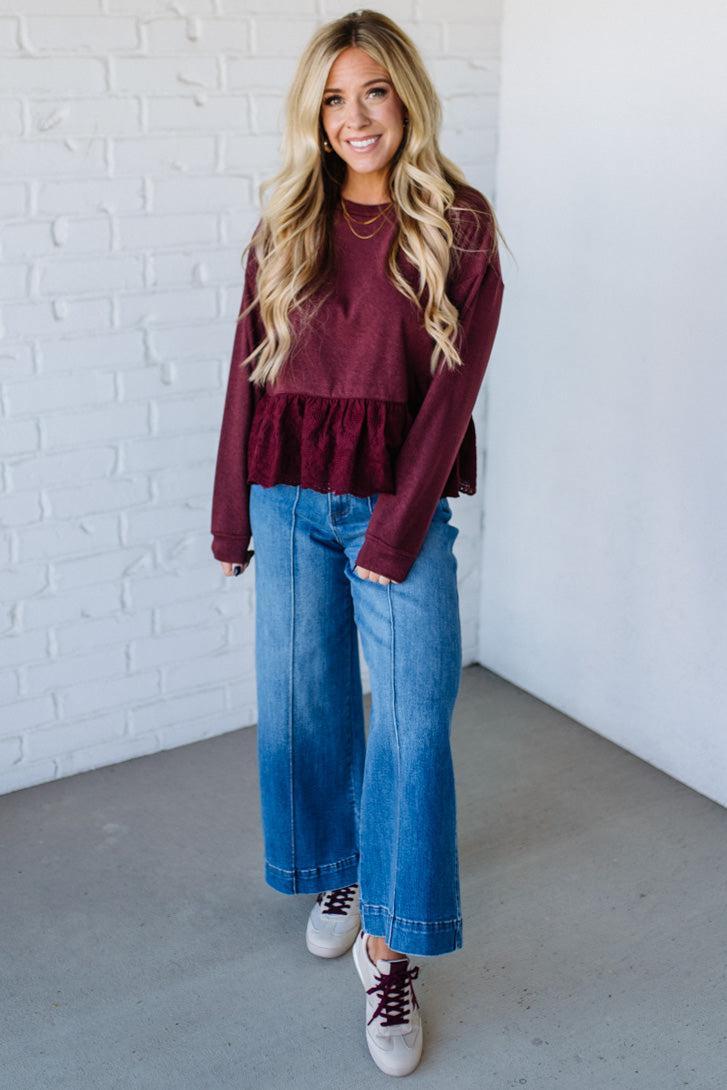 Woman wearing a burgundy ruffled top and blue jeans against a white brick wall.
