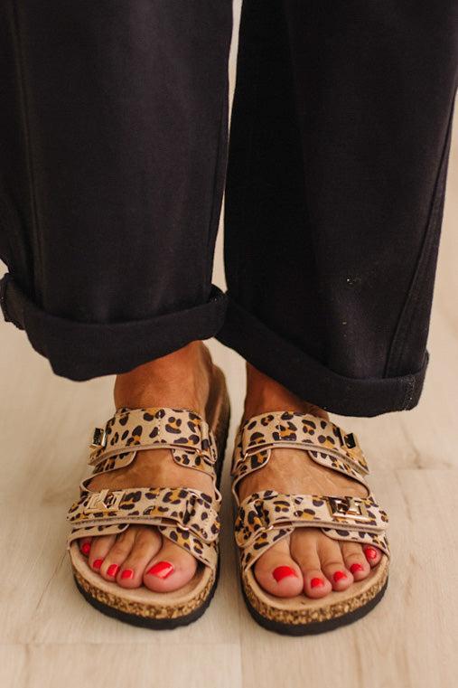 Close up of women's leopard print double strap sandals with gold buckles on feet with painted orange toenails, and black cuffed pants.