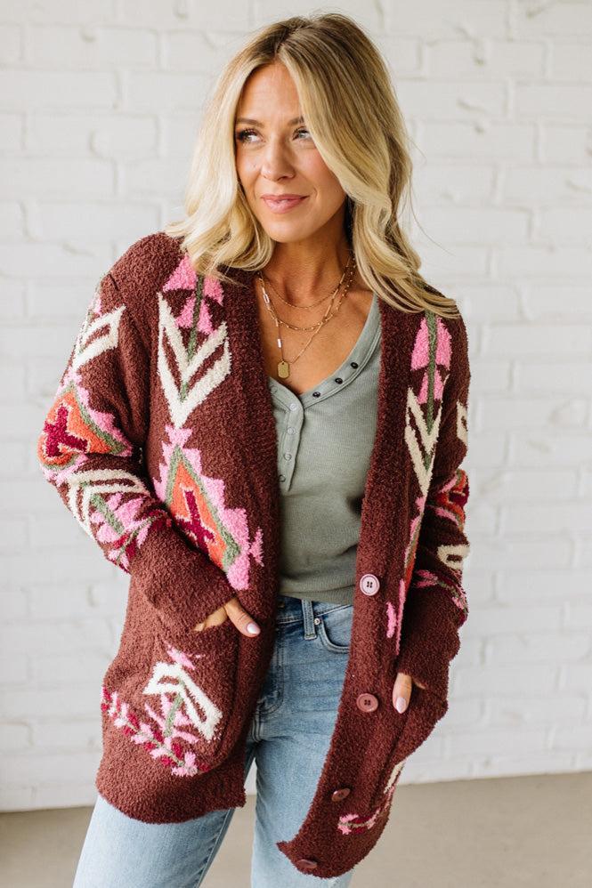 Woman wearing a patterned brown cardigan against a white brick wall.