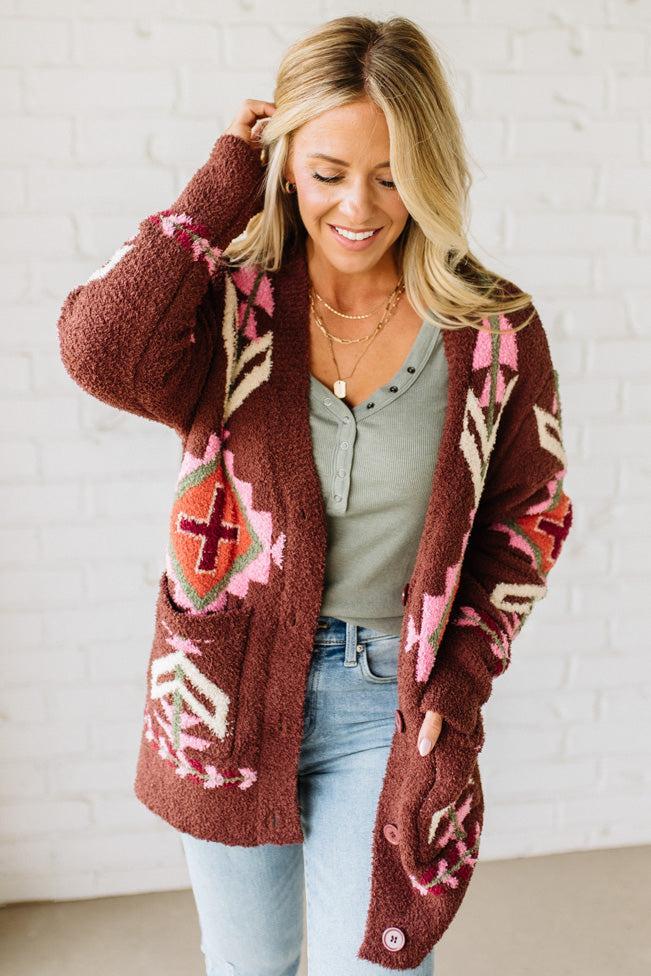 Woman wearing a patterned brown cardigan against a white brick wall.