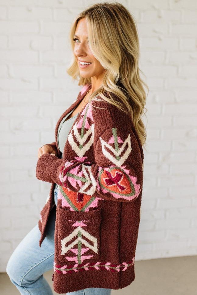 Woman wearing a brown patterned cardigan against a white brick wall.