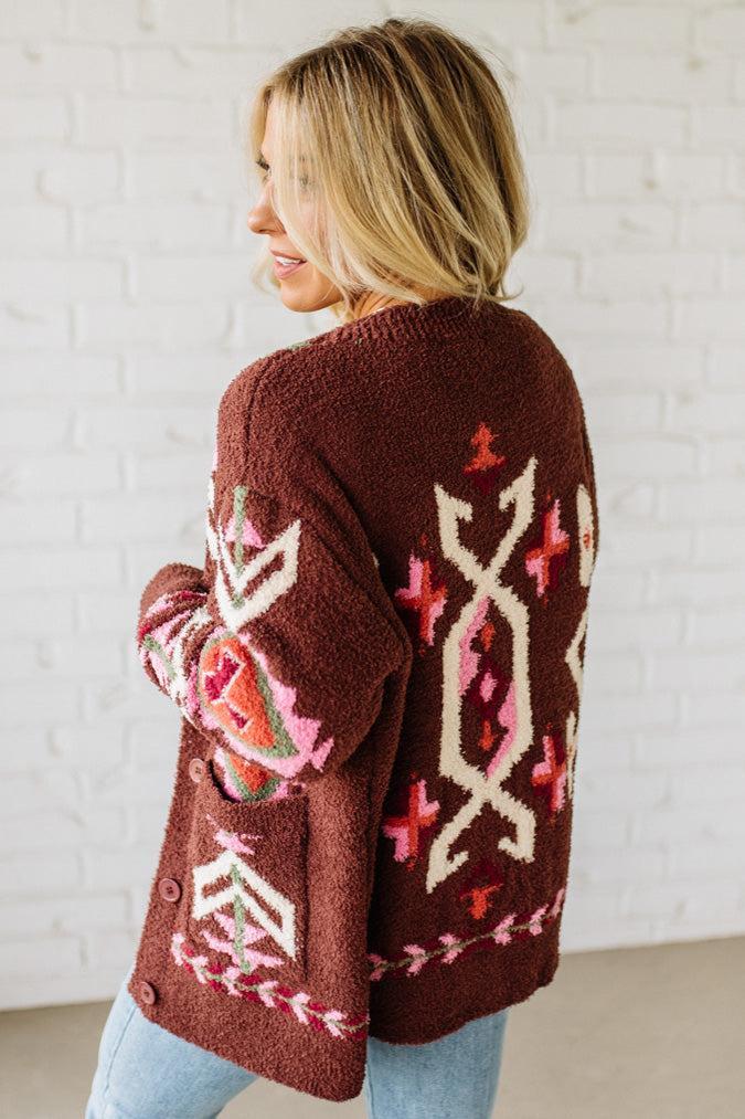 Woman wearing a brown cardigan with colorful patterns against a white brick wall.