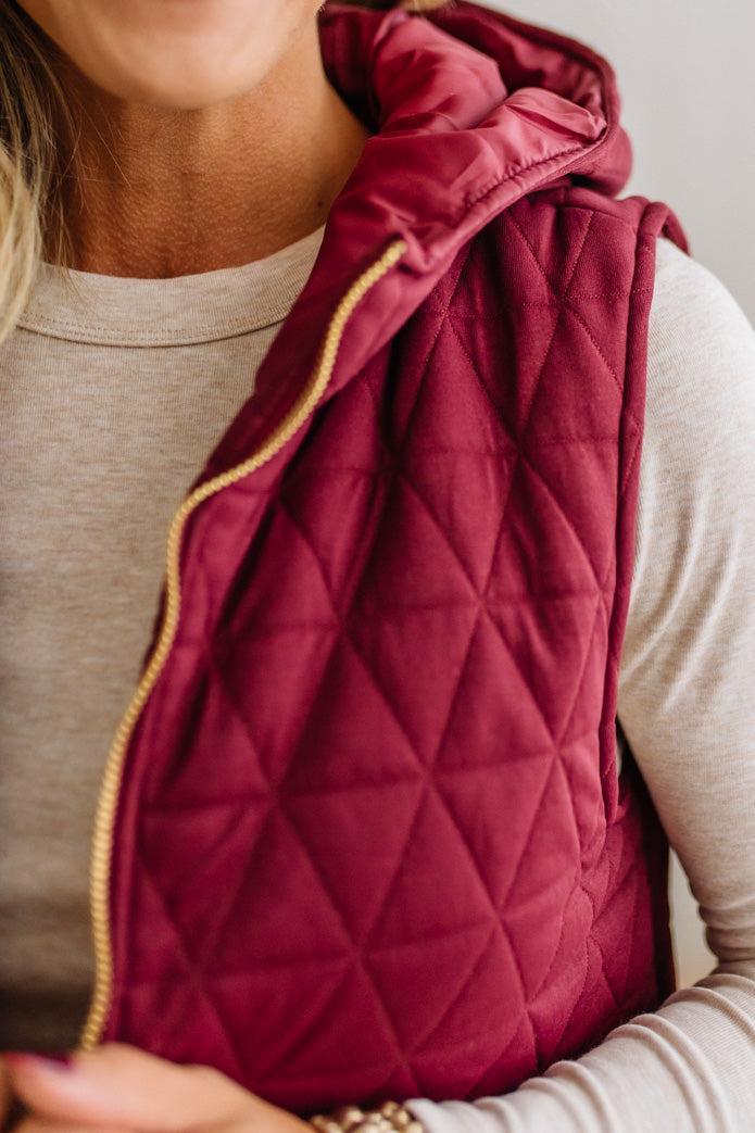 A close-up of a person wearing a quilted sleeveless puffer vest in deep burgundy with a gold zipper, layered over a light beige long-sleeve shirt.