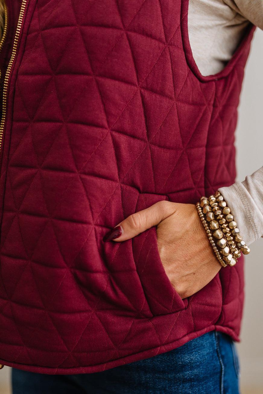 A close-up of a quilted burgundy vest with a diamond pattern, featuring a gold zipper and side pocket with a hand tucked inside, worn over a light gray top and blue jeans.