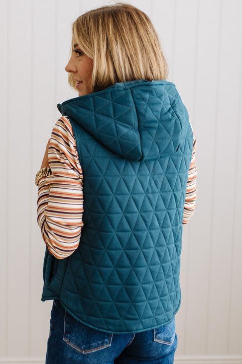 A woman shown from the back wearing a teal diamond-quilted hooded vest over a striped long-sleeve top and blue jeans, standing against a white paneled wall.