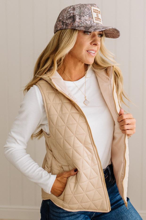 A woman wearing a beige quilted zip-front vest over a white long-sleeve shirt and blue jeans, with a camouflage baseball cap. She is standing against a light beige paneled wall, holding the vest open.