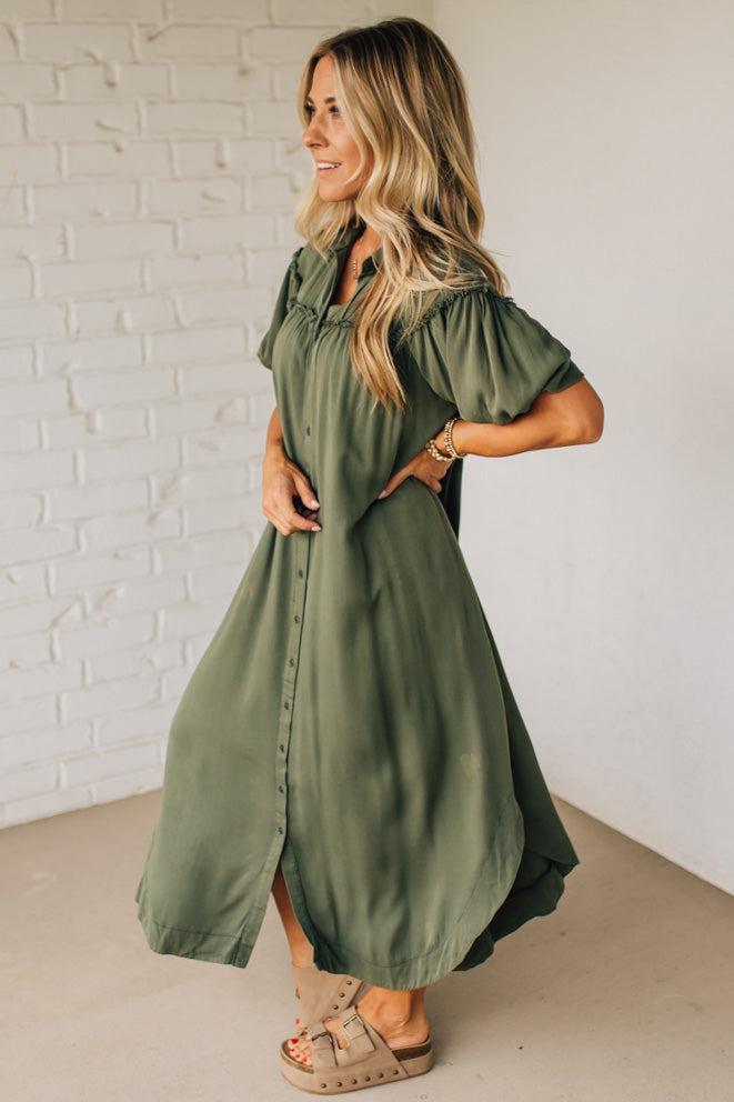 Woman wearing an olive green dress against a white brick wall.