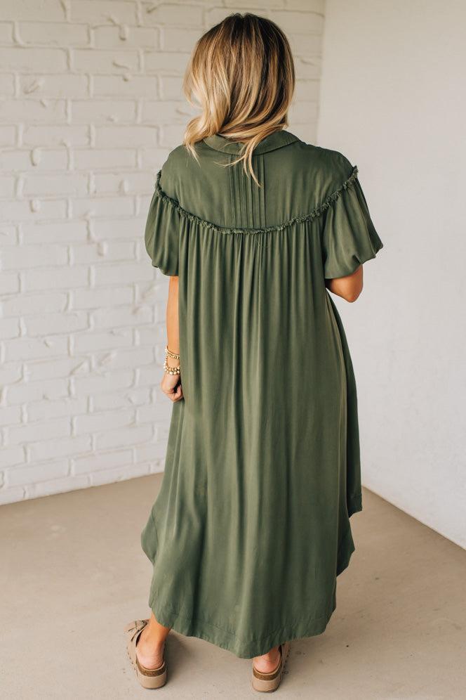 Woman wearing an olive green dress against a white brick wall.
