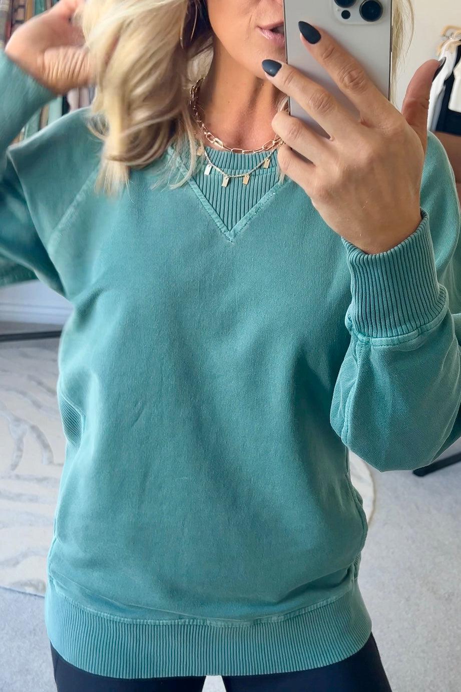 A woman wearing a teal ribbed accent pocketed pullover with dolman sleeves and a round neckline.