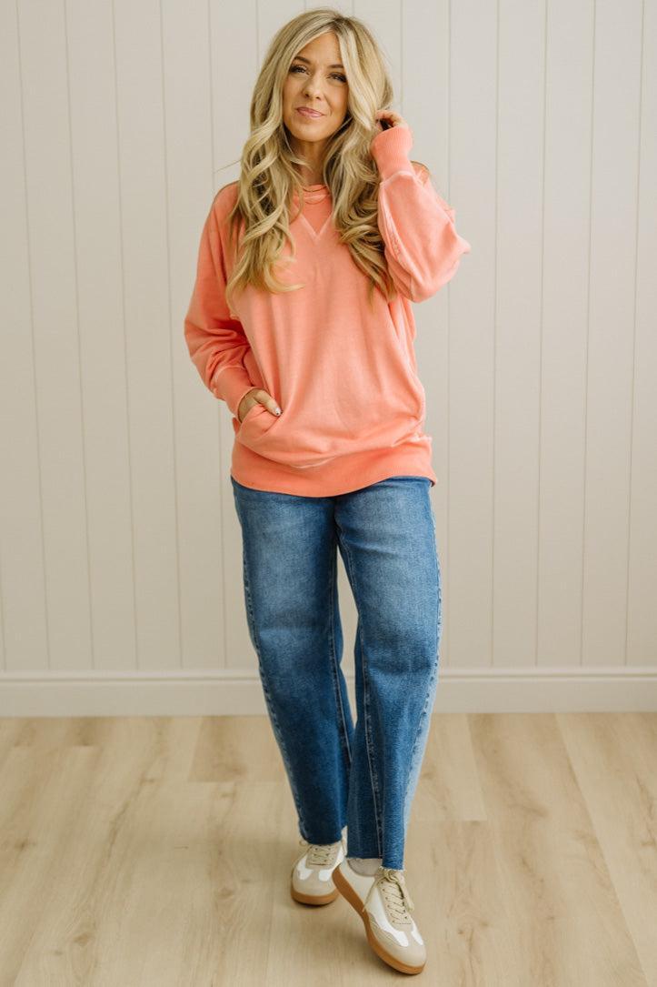 Woman wearing a pink sweatshirt and blue jeans standing against a light wooden paneled wall.