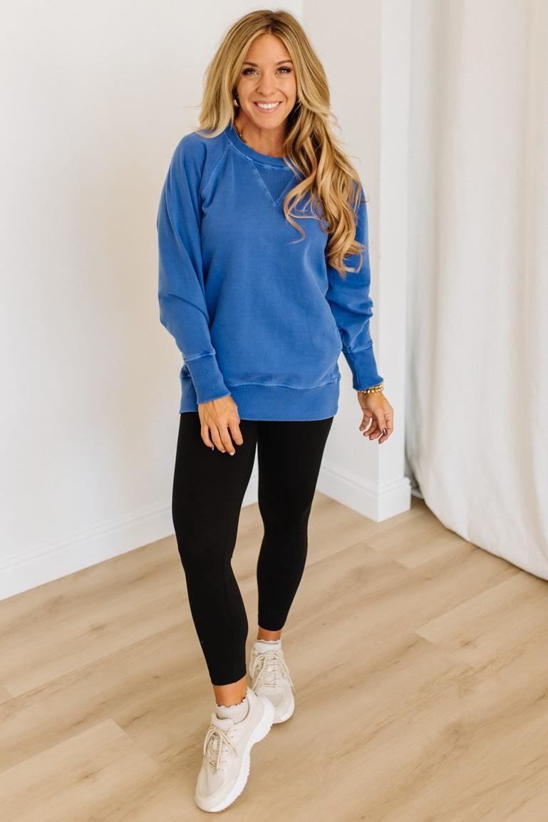 Woman wearing a blue sweatshirt, black leggings, and white sneakers in a room with light wood flooring and white walls.