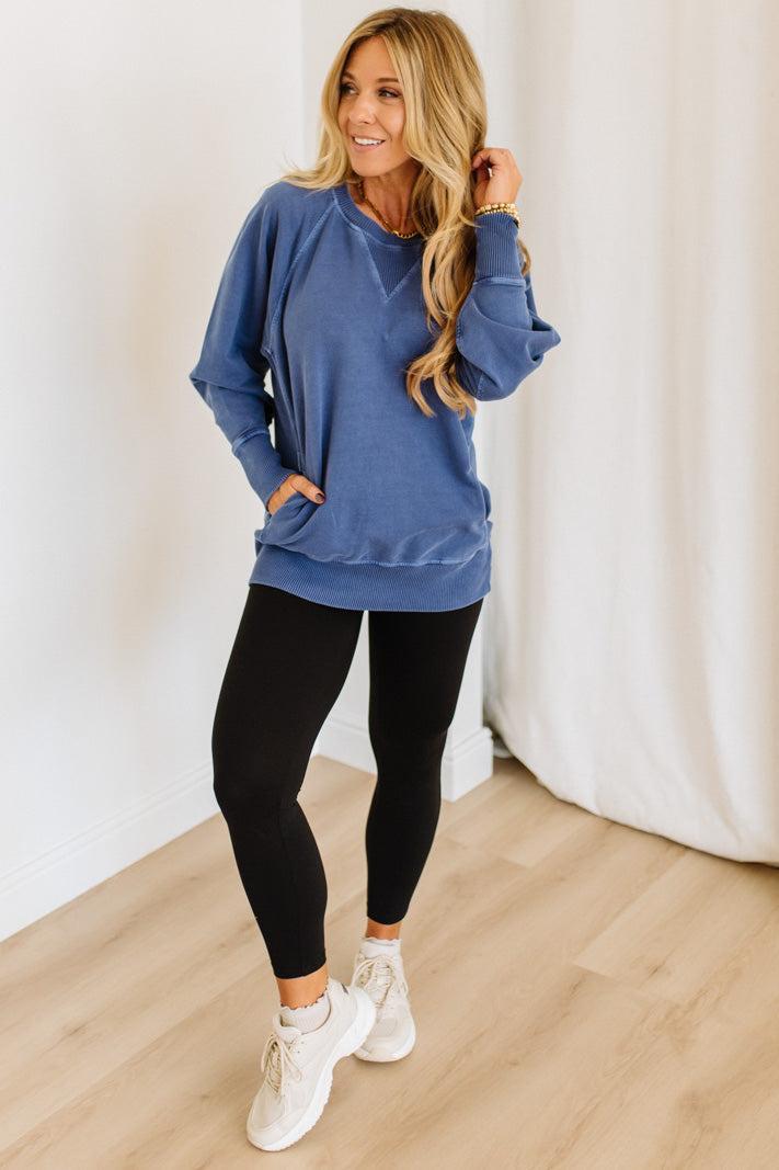Woman wearing a blue sweatshirt and black leggings standing against a white curtain.