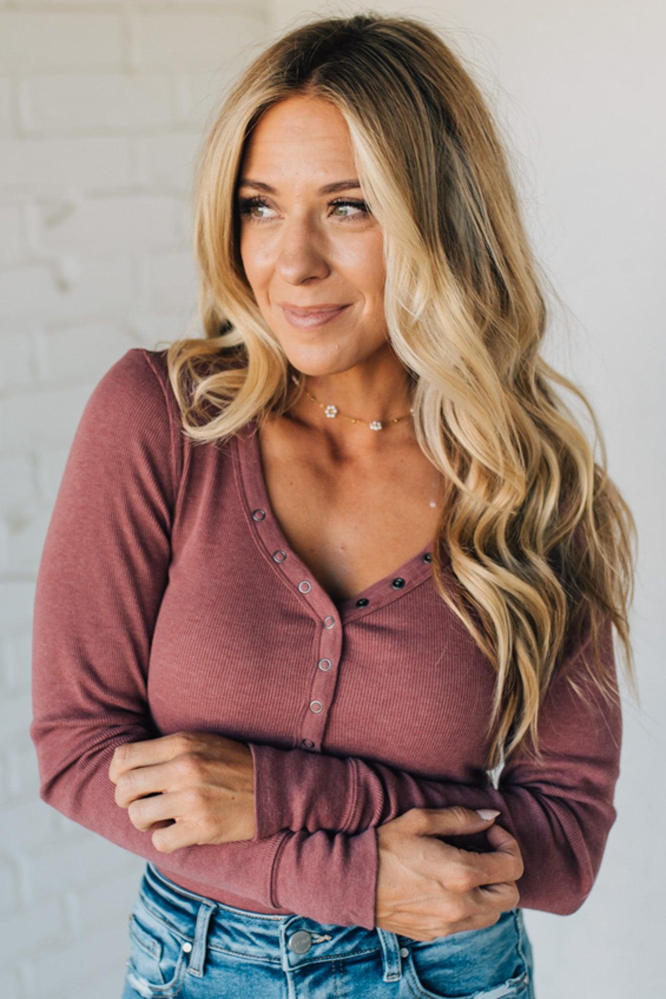 Woman wearing a mauve long-sleeve top and blue jeans against a white wall.