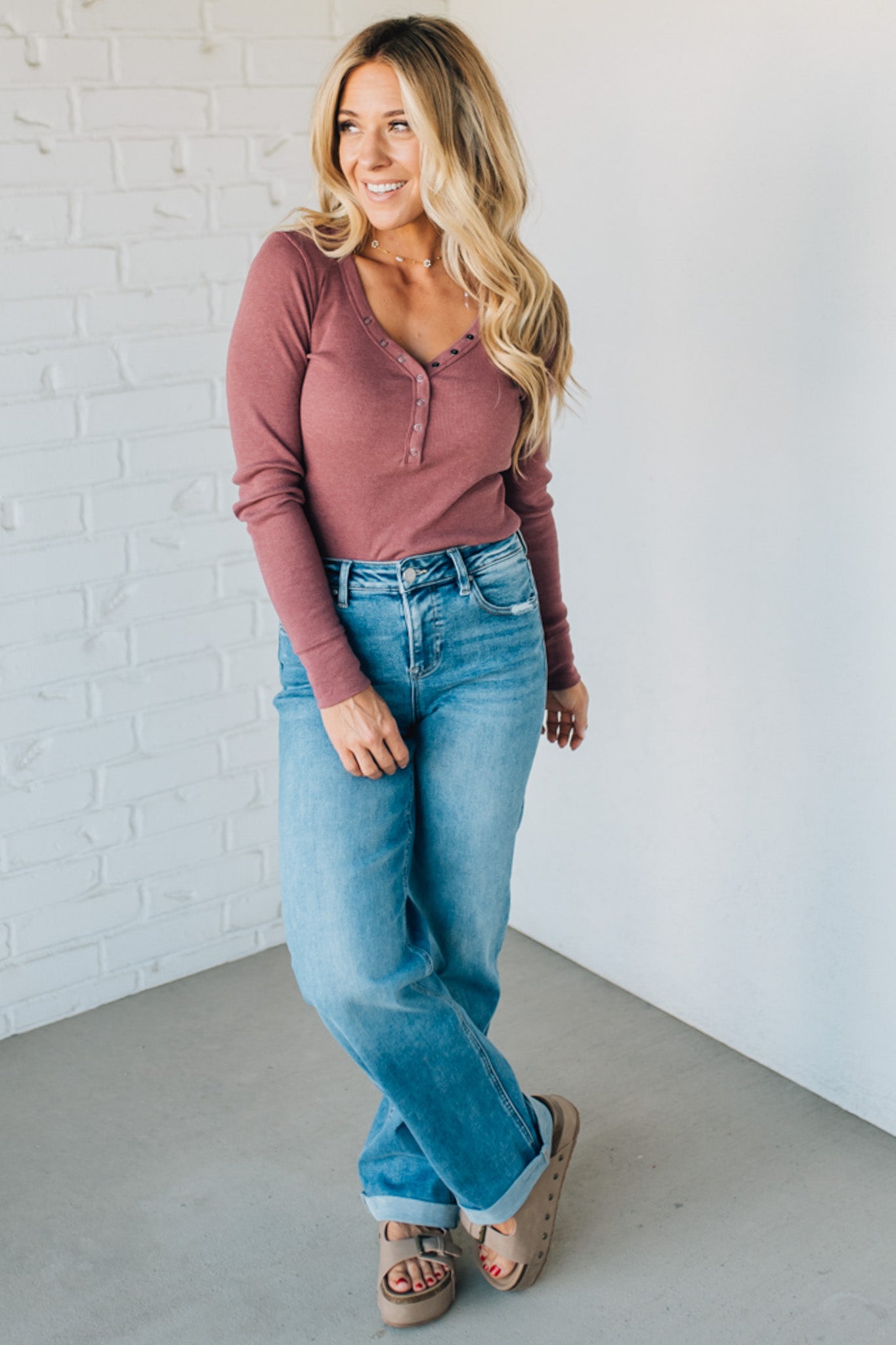 Woman wearing a pink long-sleeve top and blue jeans standing against a white brick wall.