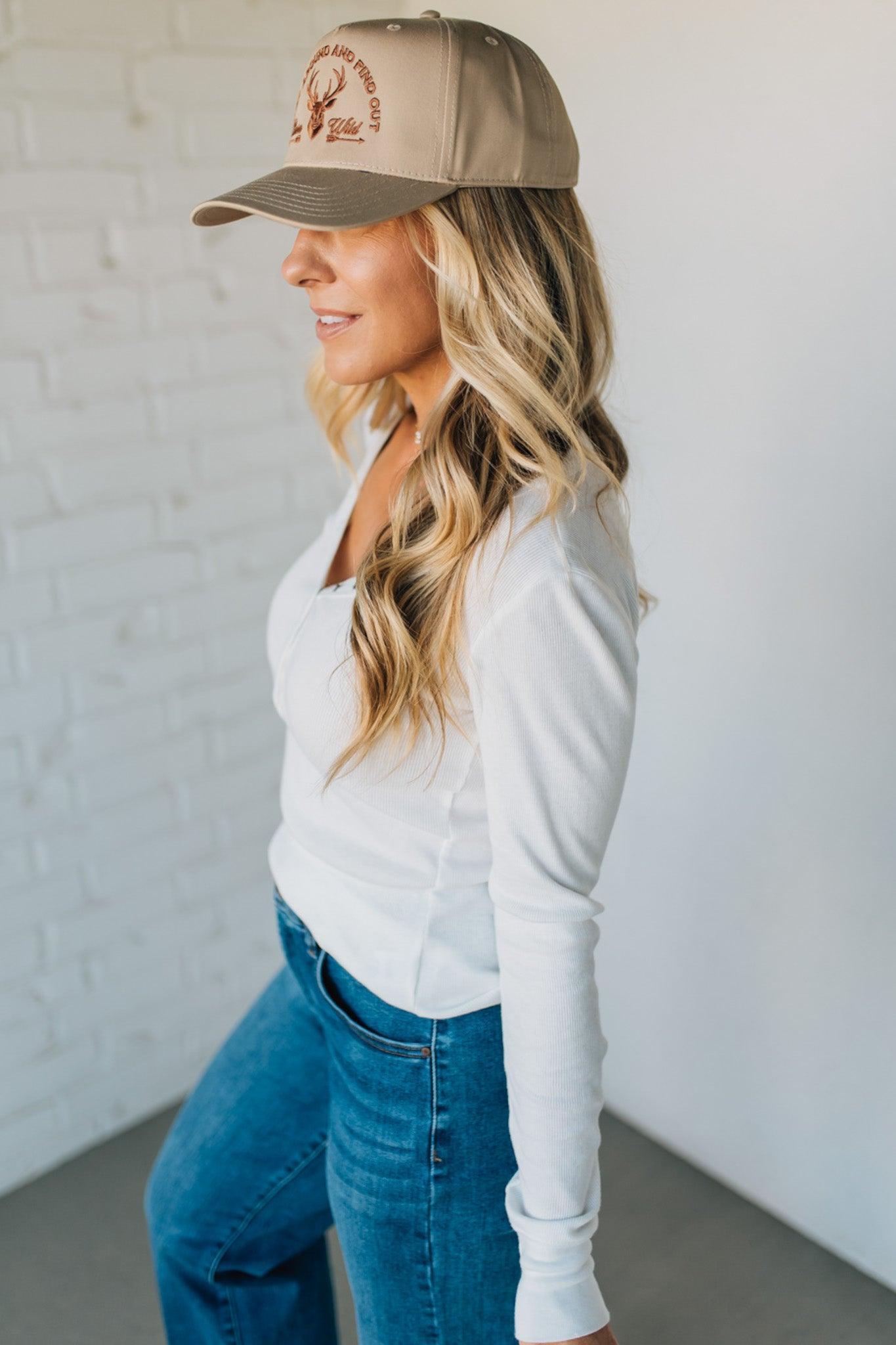 Woman wearing a beige cap with a logo, white long-sleeve shirt, and blue jeans against a white background