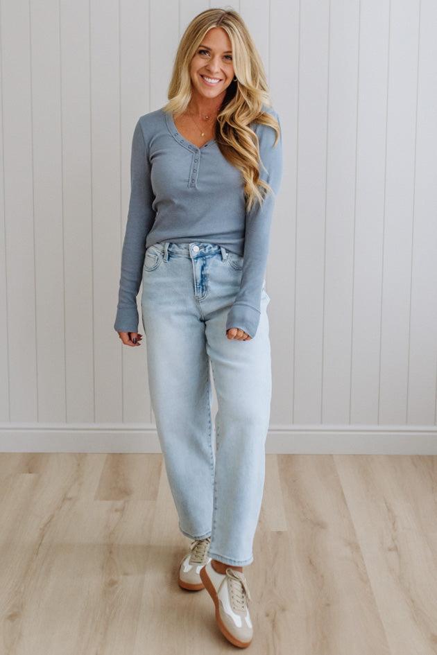 Woman wearing a blue long-sleeve top and light blue jeans standing against a white paneled wall.