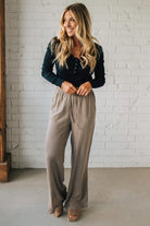 Woman wearing a black top and beige pants standing against a white brick wall.