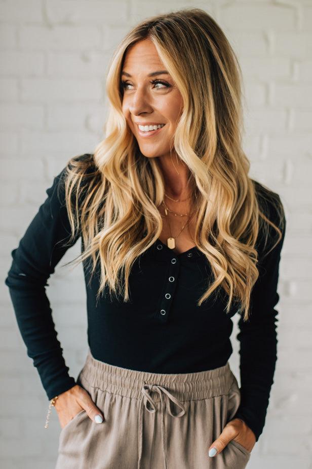 Woman wearing a black long-sleeve top and beige pants against a white brick wall.