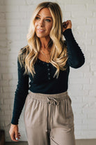 Woman wearing a black long-sleeve top and beige pants against a white brick wall.