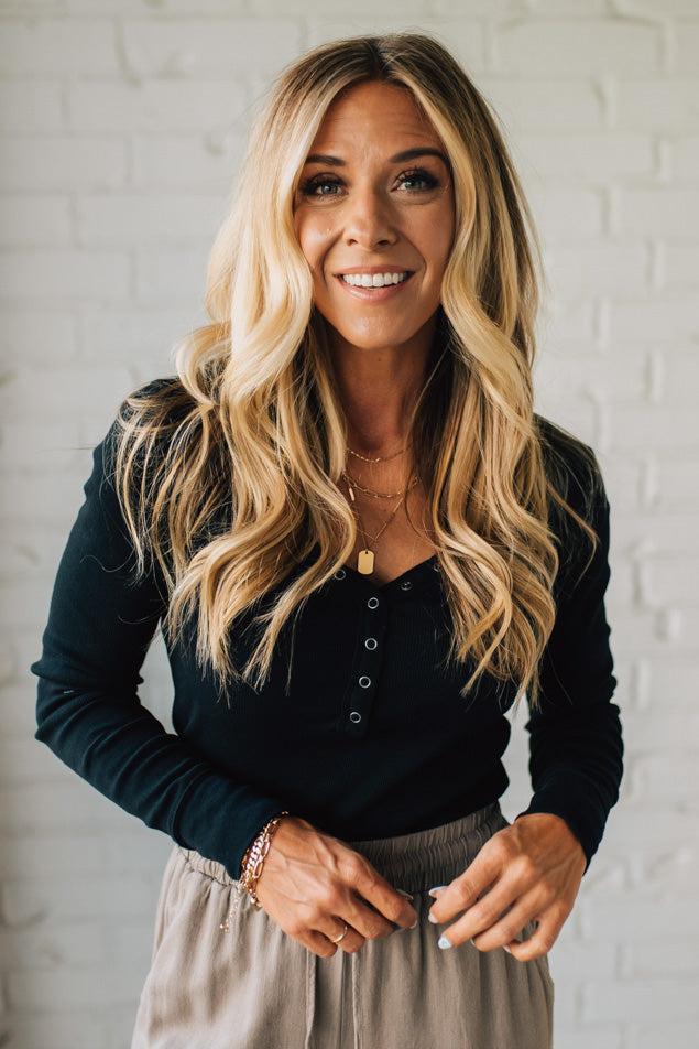 Woman with long blonde hair wearing a black long-sleeve top and beige pants against a white brick wall.