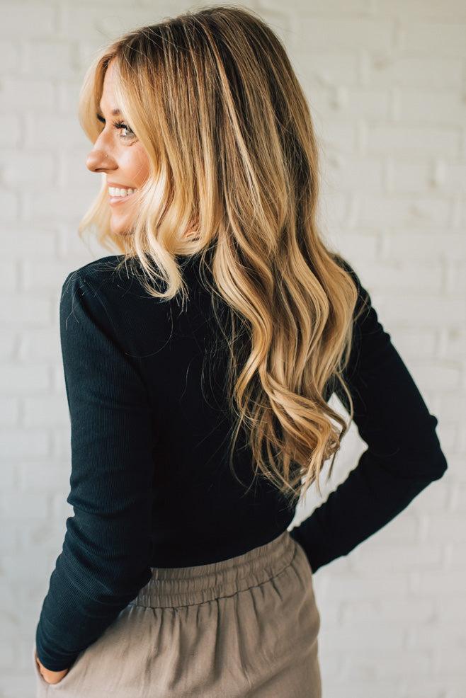 Woman wearing a black long-sleeve top and beige pants against a white brick wall.