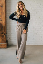 Woman wearing a black long-sleeve top and beige wide-leg pants against a white brick wall.