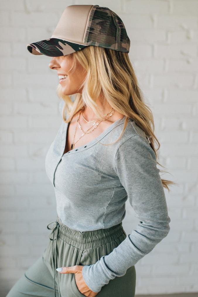 Woman wearing a gray long-sleeve top and green pants with a beige and black cap against a white brick wall.