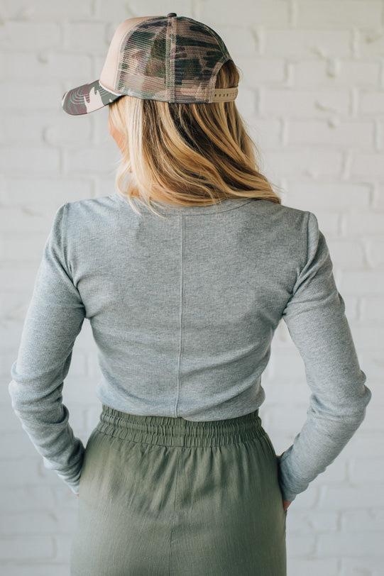 Person wearing a gray long-sleeve top and green pants with a camouflage cap, standing against a white brick wall.