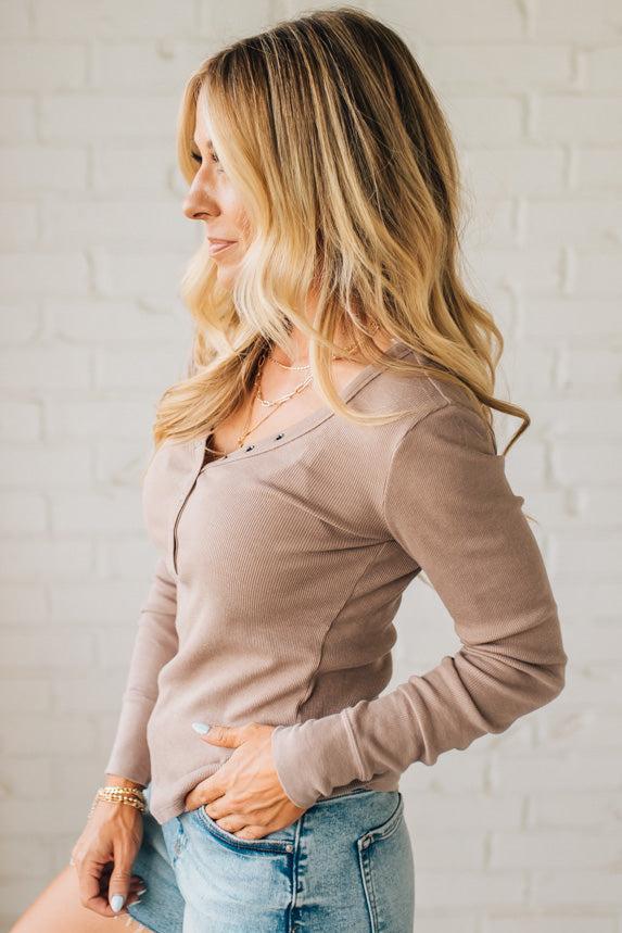 Woman wearing a beige long-sleeve top and denim shorts against a white brick wall.