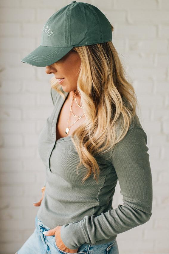 Woman wearing a green cap and long-sleeve shirt against a white brick wall.