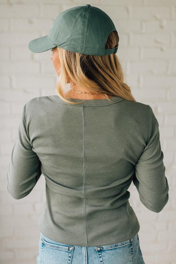 Person wearing an olive green long-sleeve top and cap, standing against a white brick wall.