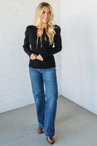 Woman wearing a black top and blue jeans against a white brick wall.