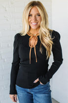Woman wearing a black sweater with a tie detail and blue jeans against a white brick wall.