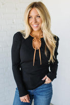 Woman wearing a black sweater with a brown tie and blue jeans against a white brick wall.