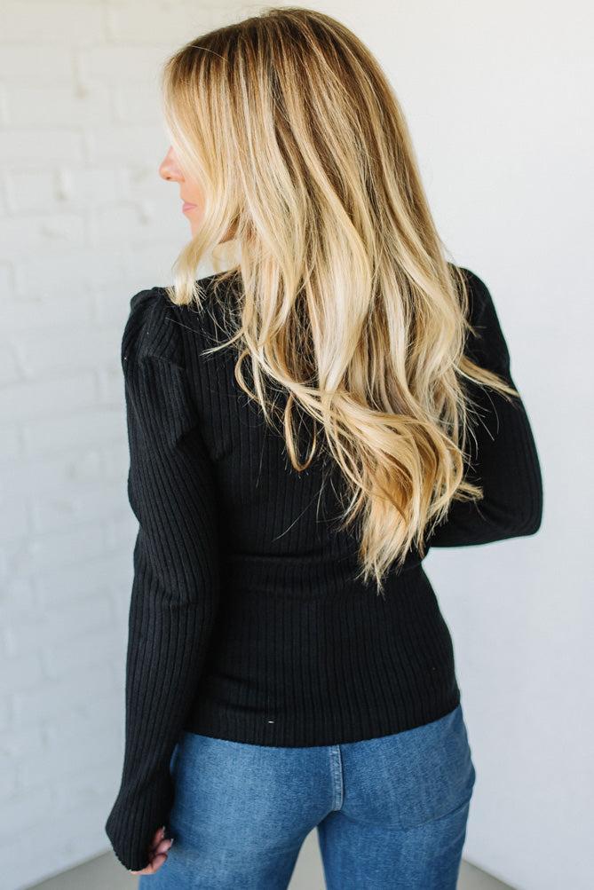 Woman wearing a black ribbed sweater and blue jeans against a white wall