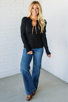 Woman wearing a black sweater with a brown bow and blue jeans against a white brick wall.