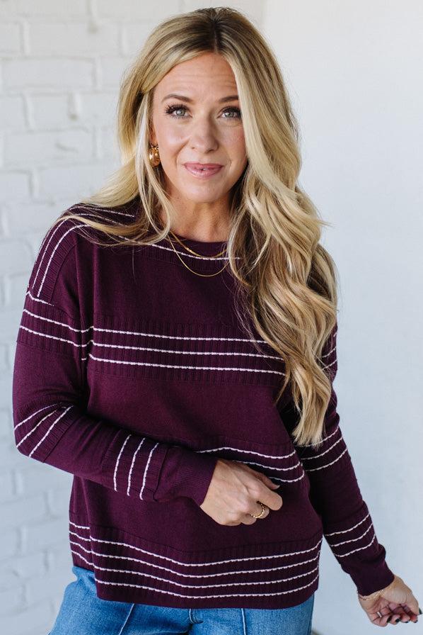 Woman wearing a maroon sweater with white stripes against a white brick wall.