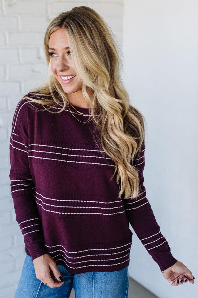 Woman wearing a maroon sweater with white stripes against a white brick wall.