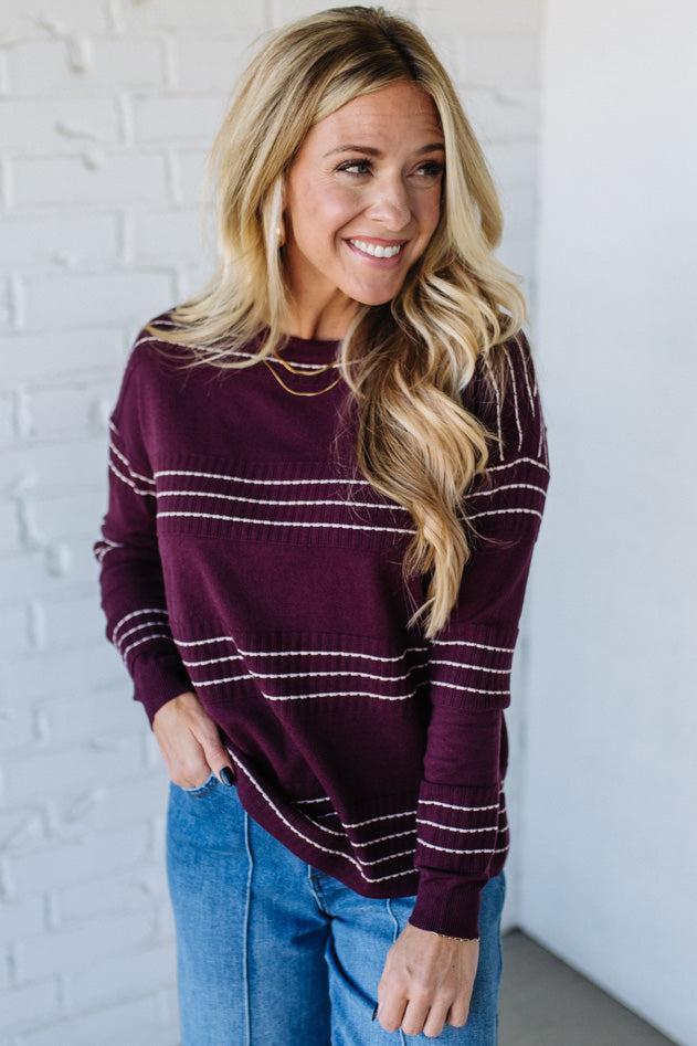Woman wearing a maroon sweater with white stripes against a white brick wall.