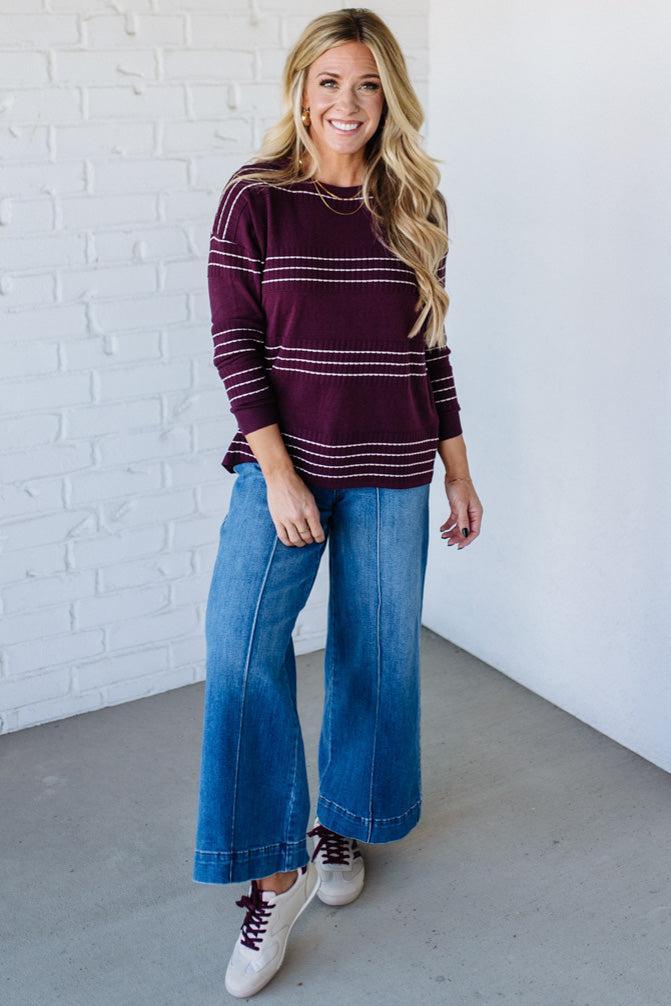 Woman wearing a striped sweater and blue jeans against a white brick wall.
