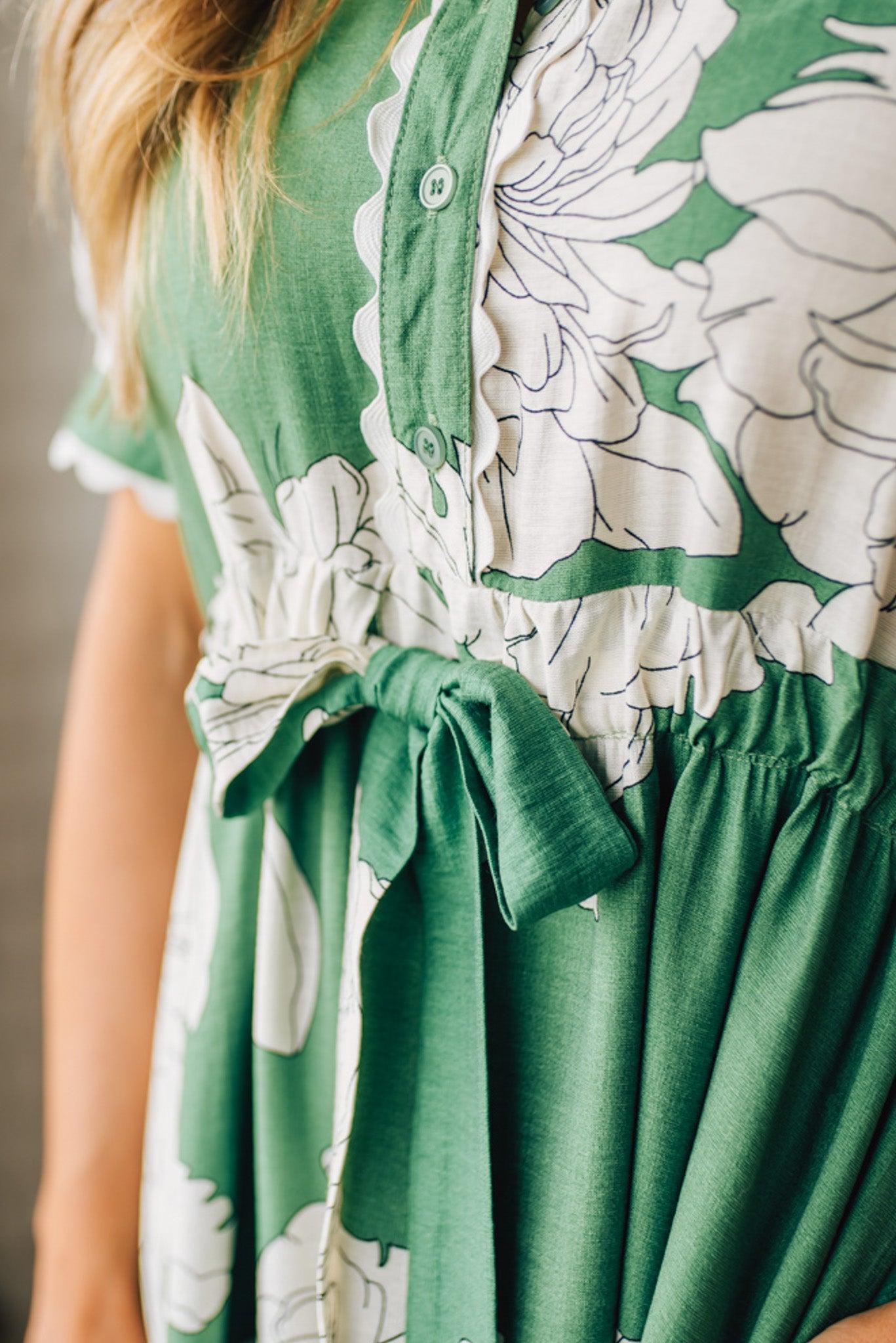 Blonde woman wearing a flower print midi dress with collared neck features buttoned closure, belted waistline, side pockets, puffed short sleeves, tiered layer skirt, and trimming along sleeves and neck.