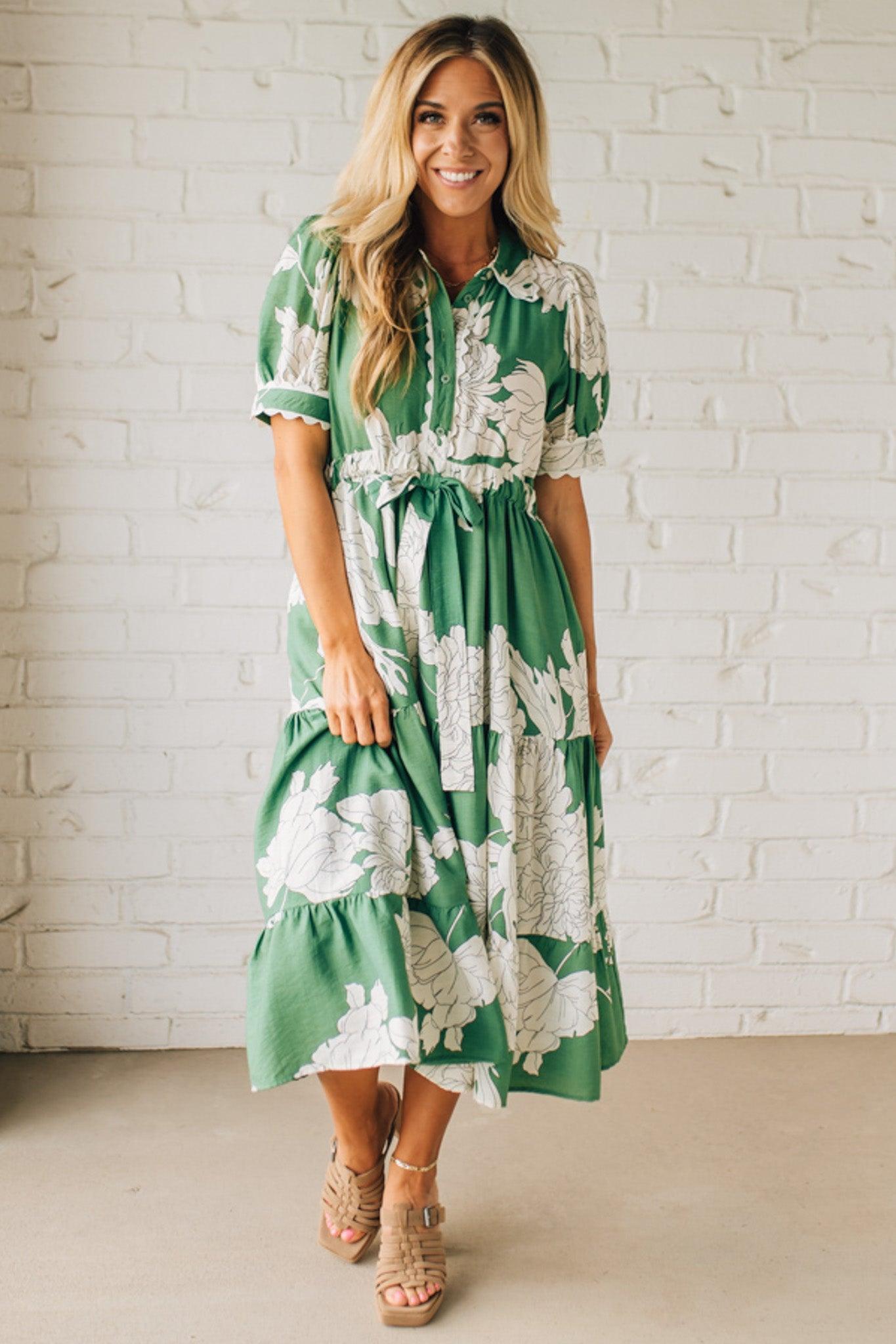 Blonde woman wearing a flower print midi dress with collared neck features buttoned closure, belted waistline, side pockets, puffed short sleeves, tiered layer skirt, and trimming along sleeves and neck.