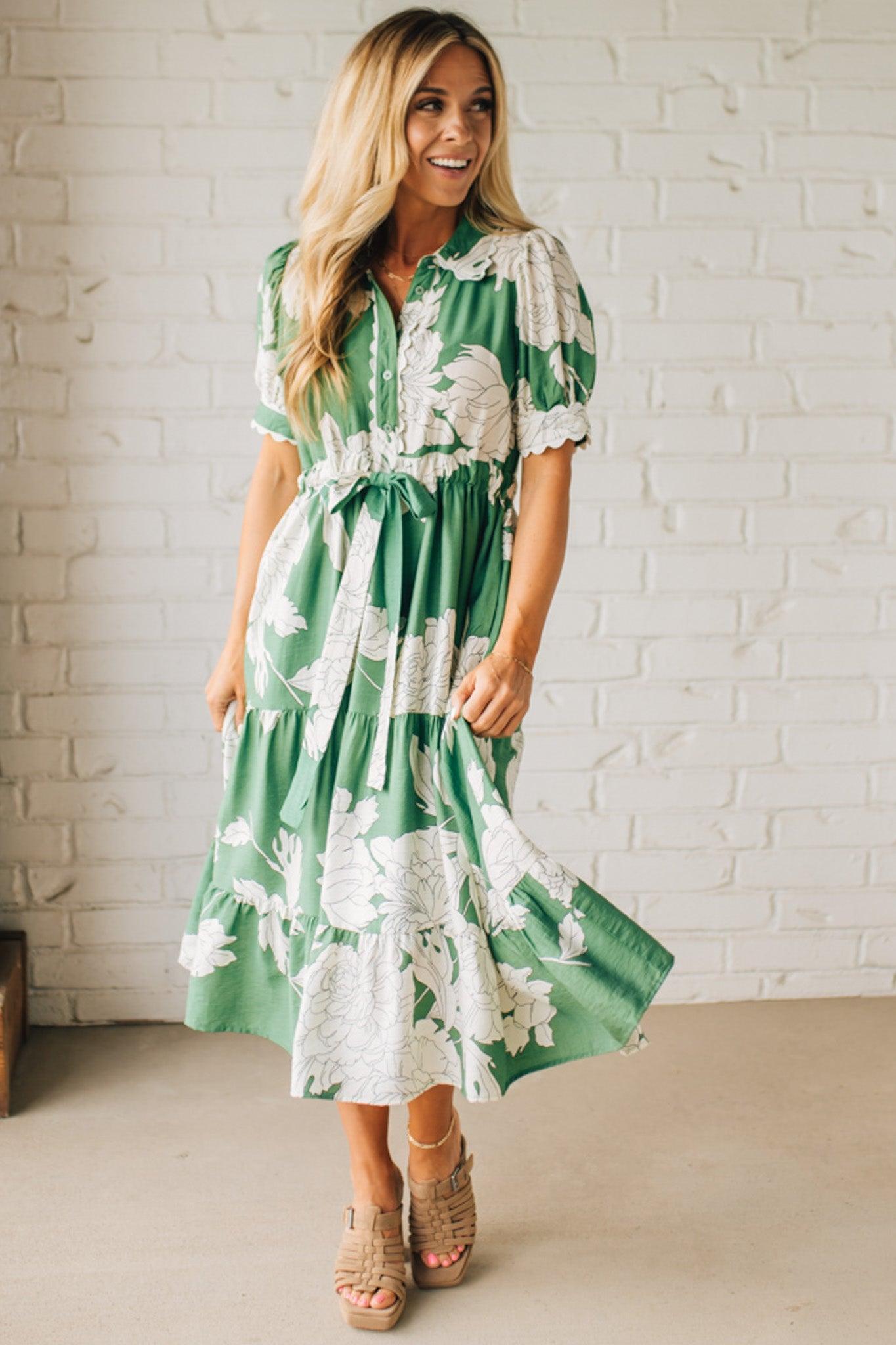 Blonde woman wearing a flower print midi dress with collared neck features buttoned closure, belted waistline, side pockets, puffed short sleeves, tiered layer skirt, and trimming along sleeves and neck.